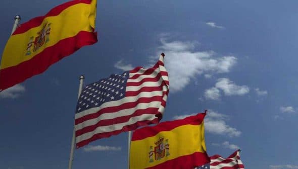 Spanish flags and an American flag waving in the wind against a blue sky with clouds. - Olive Oil Times