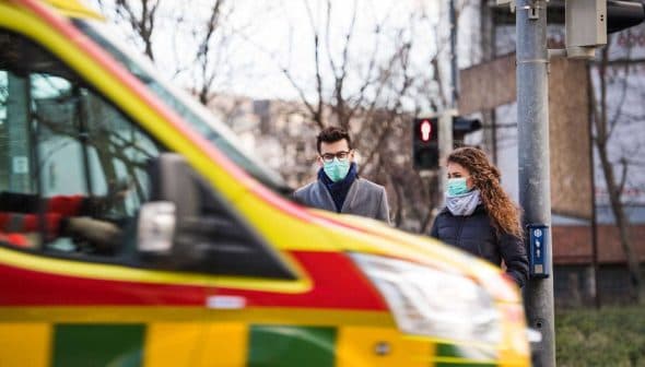 Two individuals wearing masks waiting at a crosswalk as an ambulance drives by in the foreground. - Olive Oil Times