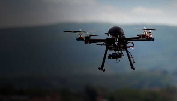 A quadcopter drone flying in the air with a blurred background of mountains. - Olive Oil Times