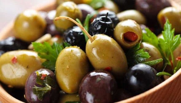 A wooden bowl filled with a variety of green and black olives, garnished with herbs. - Olive Oil Times