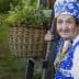 Elderly woman wearing traditional blue and white attire with a floral headscarf, holding a wooden post beside a basket of flowers. - Olive Oil Times