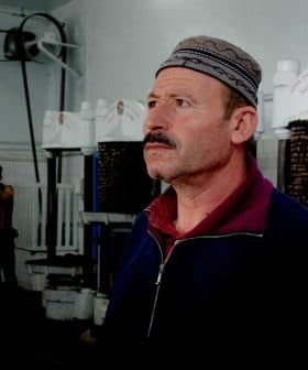 Man wearing a traditional hat standing inside an oil pressing facility with machinery in the background. - Olive Oil Times