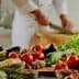A variety of fresh vegetables, herbs, and cooking ingredients arranged on a kitchen counter with a chef preparing food in the background. - Olive Oil Times