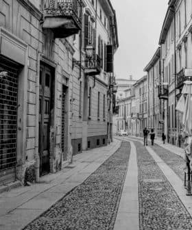 Black and white image of a narrow cobblestone street with buildings and pedestrians. - Olive Oil Times