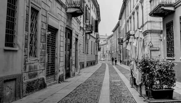 Black and white image of a narrow cobblestone street with buildings and pedestrians. - Olive Oil Times
