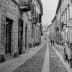 Black and white image of a narrow cobblestone street with buildings and pedestrians. - Olive Oil Times