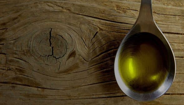 Metal spoon filled with olive oil resting on a textured wooden surface. - Olive Oil Times