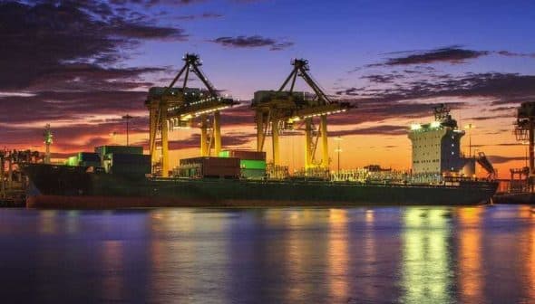 Cargo ship docked at a port with cranes and containers during a sunset. - Olive Oil Times