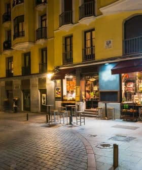 Street view of a yellow building featuring a café with outdoor seating and illuminated signage at night. - Olive Oil Times
