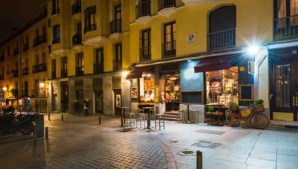 Street view of a yellow building featuring a café with outdoor seating and illuminated signage at night. - Olive Oil Times