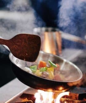 Chef using a wooden spatula to stir colorful vegetables in a frying pan over a flame. - Olive Oil Times