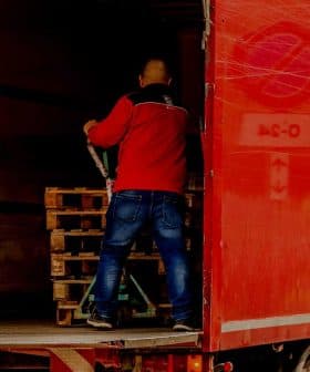 A man in a red jacket loading wooden pallets into the back of a red delivery truck. - Olive Oil Times