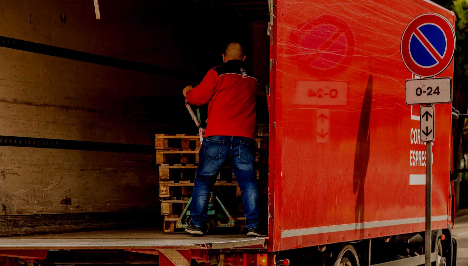 A man in a red jacket loading wooden pallets into the back of a red delivery truck. - Olive Oil Times