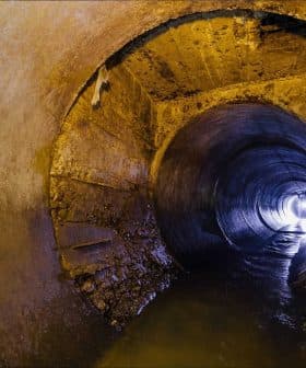 Interior view of a large concrete drainage pipe with water flowing through it and light at the end. - Olive Oil Times