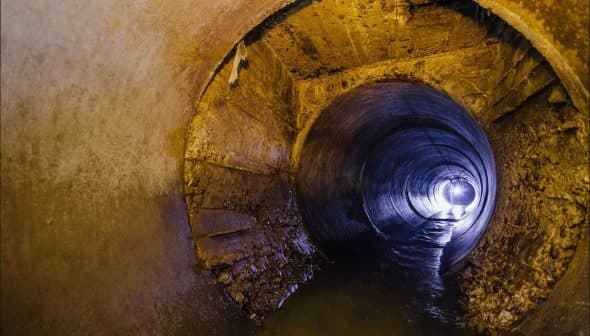 Interior view of a large concrete drainage pipe with water flowing through it and light at the end. - Olive Oil Times