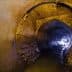 Interior view of a large concrete drainage pipe with water flowing through it and light at the end. - Olive Oil Times