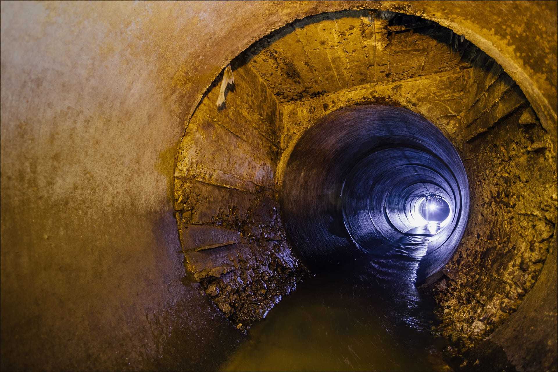 Interior view of a large concrete drainage pipe with water flowing through it and light at the end. - Olive Oil Times