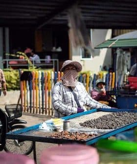 A street vendor wearing a mask and hat stands behind a table with various snacks for sale. - Olive Oil Times