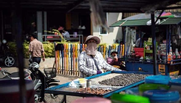A street vendor wearing a mask and hat stands behind a table with various snacks for sale. - Olive Oil Times
