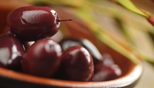 A close-up view of Kalamata olives in a bowl, showcasing their dark purple color and glossy surface. - Olive Oil Times