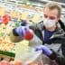 Man in a mask and gloves placing an apple into a plastic bag in a grocery store. - Olive Oil Times
