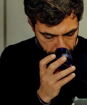 A man with dark hair and a beard holding a blue glass cup close to his face. - Olive Oil Times