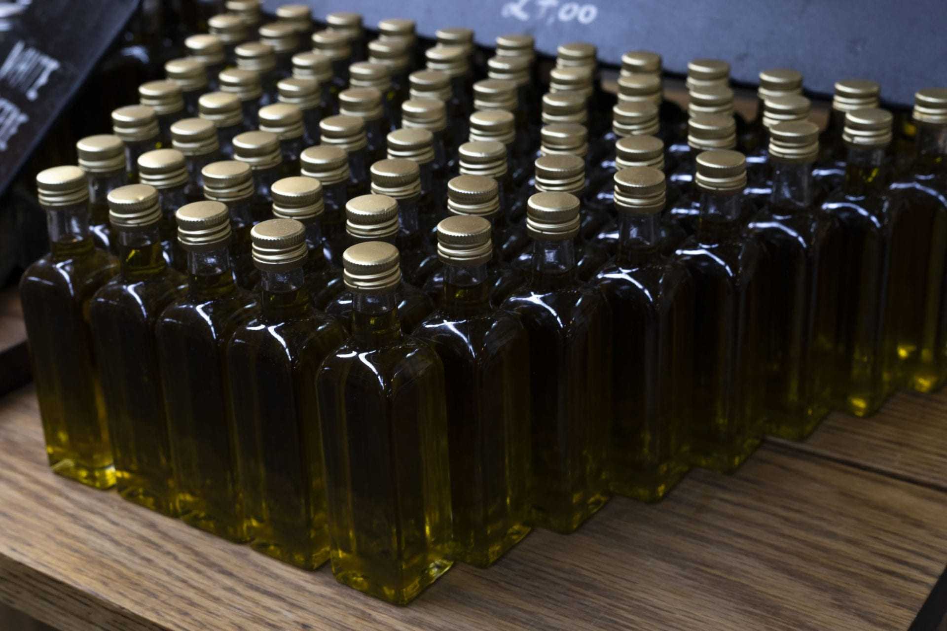 Row of small glass bottles filled with olive oil, each with a gold cap, arranged neatly on a wooden surface. - Olive Oil Times