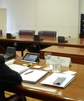 Man attending a virtual meeting with multiple participants displayed on a screen in a conference room. - Olive Oil Times