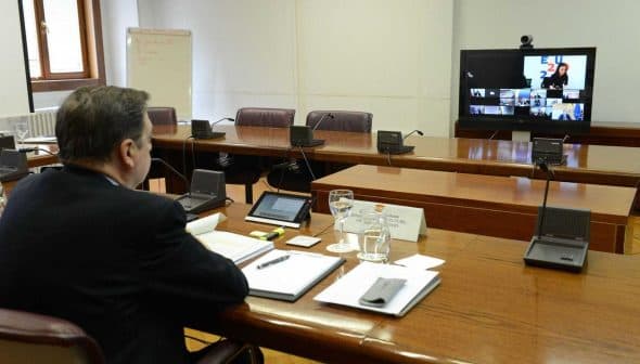 Man attending a virtual meeting with multiple participants displayed on a screen in a conference room. - Olive Oil Times