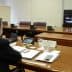 Man attending a virtual meeting with multiple participants displayed on a screen in a conference room. - Olive Oil Times