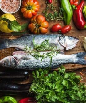 Two whole fish placed on a wooden board surrounded by various vegetables and herbs. - Olive Oil Times
