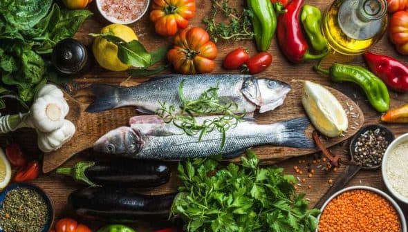 Two whole fish placed on a wooden board surrounded by various vegetables and herbs. - Olive Oil Times
