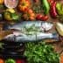Two whole fish placed on a wooden board surrounded by various vegetables and herbs. - Olive Oil Times