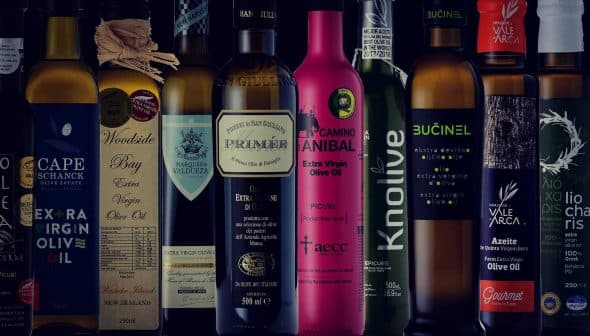 A collection of olive oil bottles with different labels and designs arranged in a row. - Olive Oil Times