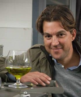 Man smiling while leaning on a machine with a glass of green olive oil in front of him. - Olive Oil Times