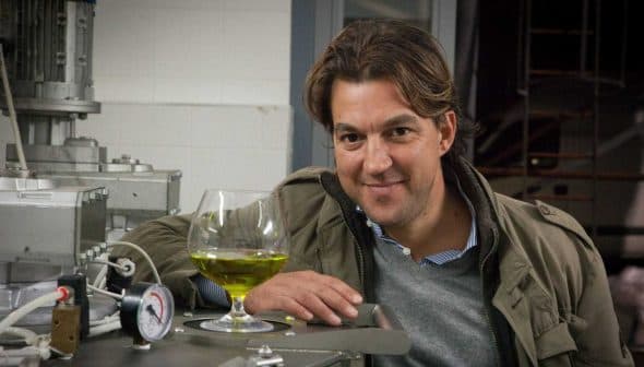 Man smiling while leaning on a machine with a glass of green olive oil in front of him. - Olive Oil Times