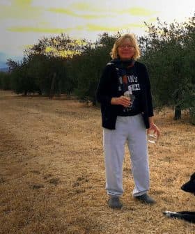A woman holding a glass and standing next to two dogs in an olive grove. - Olive Oil Times