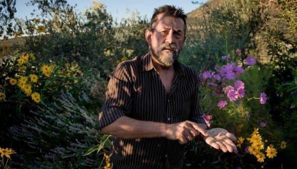 Man with a beard holding soil in his hand while standing among colorful flowers. - Olive Oil Times