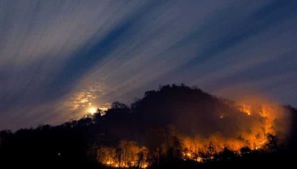 A mountain landscape with visible flames and smoke under a night sky with clouds and a full moon. - Olive Oil Times