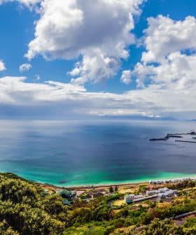 Panoramic view of a coastal landscape featuring a beach, ocean, and distant mountains under a cloudy sky. - Olive Oil Times