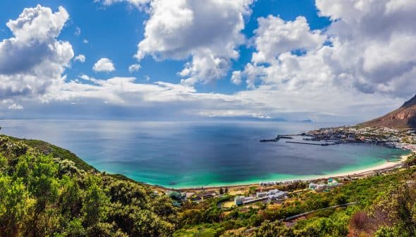 Panoramic view of a coastal landscape featuring a beach, ocean, and distant mountains under a cloudy sky. - Olive Oil Times