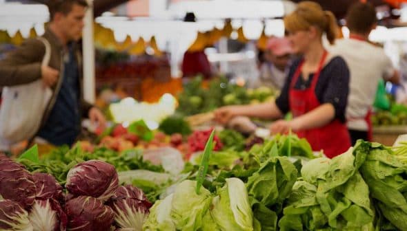 Fresh vegetables including lettuce and radicchio displayed at a market stall with shoppers in the background. - Olive Oil Times