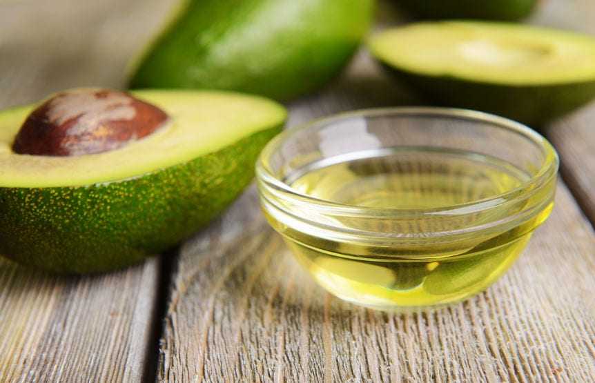Sliced avocado with a pit next to a small glass bowl of avocado oil on a wooden surface. - Olive Oil Times