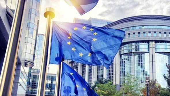 European Union flags waving in front of a modern building with glass architecture. - Olive Oil Times