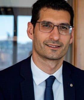 Man wearing glasses and a suit, seated in front of a wooden cabinet with books and decorative items. - Olive Oil Times