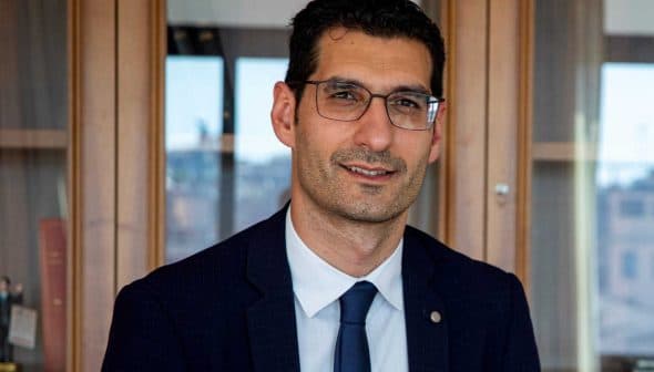 Man wearing glasses and a suit, seated in front of a wooden cabinet with books and decorative items. - Olive Oil Times