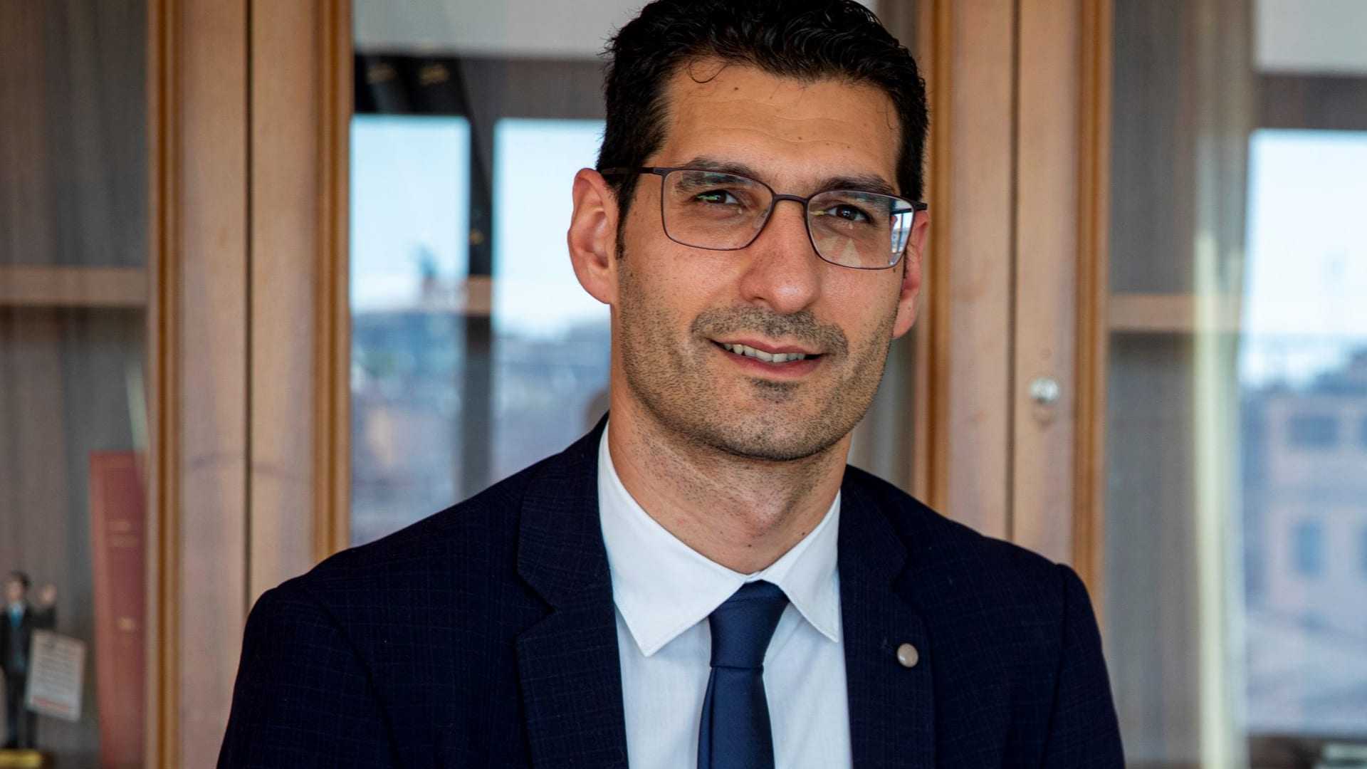 Man wearing glasses and a suit, seated in front of a wooden cabinet with books and decorative items. - Olive Oil Times