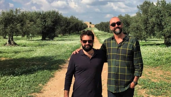Two men posing together in an olive grove with trees and grass in the background. - Olive Oil Times