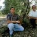Two men kneeling beside an olive tree in an olive grove, both smiling at the camera. - Olive Oil Times