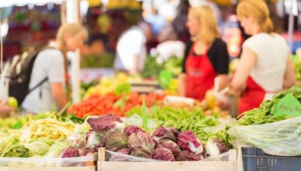 A variety of fresh vegetables displayed at a market stand with shoppers in the background. - Olive Oil Times